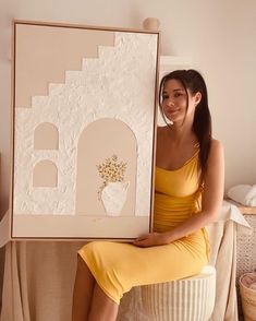 a woman sitting on a stool holding up a large piece of art that looks like a house