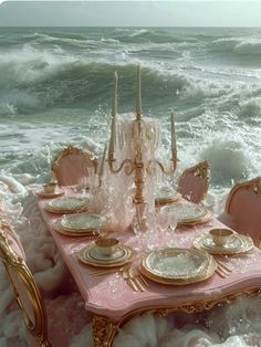 a pink table set with gold plates and candles on top of the ocean wave covered beach