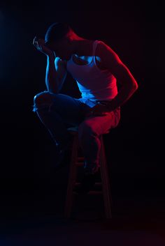 a man sitting on a stool in the dark with his hands to his face and looking down