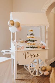 a white cart with some gold and white balloons on it's side, filled with cupcakes