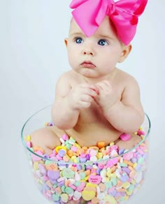 a baby sitting in a bowl filled with candies and wearing a big pink bow