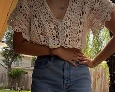 a woman standing in front of a tree with her hands on her hips, wearing jeans and a crocheted top