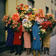 four women standing in front of a building with flowers on their heads and one holding onto the back of another woman's head