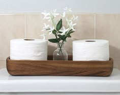 two rolls of toilet paper sitting on top of a wooden tray next to a vase with flowers