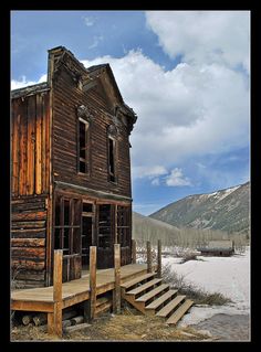 an old wooden house sitting on top of a snow covered field next to a mountain