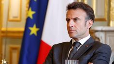 a man in a suit and tie sitting at a table with two flags behind him