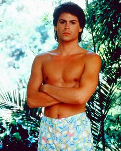 a man with his arms crossed standing in front of some plants and trees, wearing swim trunks