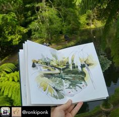 a person holding up an open book in front of some trees and water with people walking on the other side