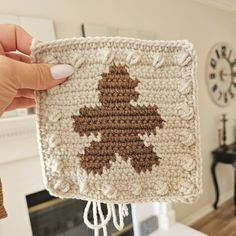 a person holding up a crocheted square with a tree on it in front of a fireplace