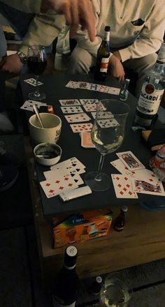 two men sitting at a table with cards and wine glasses on it, one holding a bottle