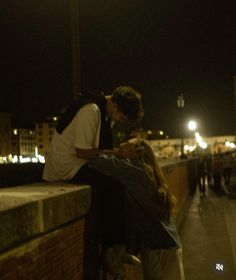 a man and woman kissing on the side of a bridge at night with city lights in the background