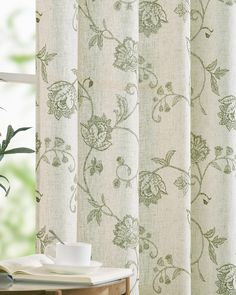 an open book on a table next to a window with curtains and flowers in it