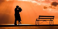 a man and woman standing next to each other in front of a bench at sunset