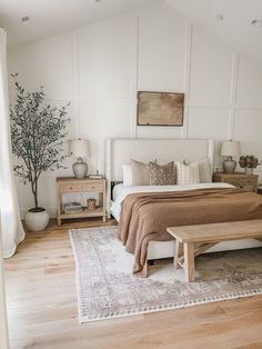 a bedroom with white walls and wood flooring has a large bed, two nightstands, and a bench in the corner