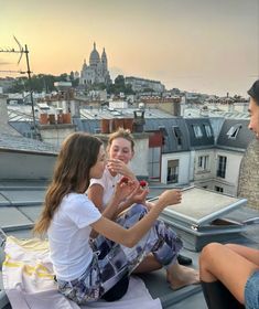 three young women sitting on the roof of a building talking to each other and drinking wine