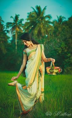 a beautiful woman in a sari holding a pot with flowers on her feet and walking through the grass