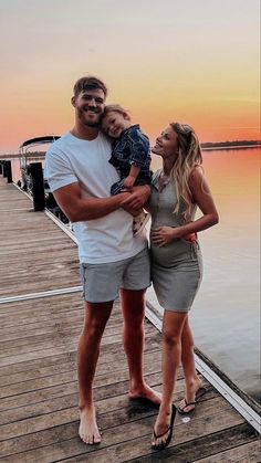 a man and woman standing on a dock with a baby in their arms at sunset