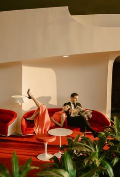 a man and woman sitting on a red couch in a room with white walls, plants and floor to ceiling windows