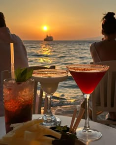 two cocktails sitting on top of a white table next to the ocean at sunset