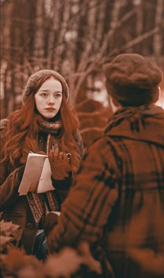 a woman with red hair standing next to another person in the woods holding a book