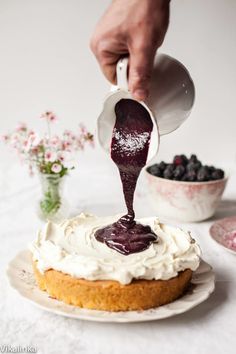 someone pouring icing onto a cake with blueberries in the bowl next to it