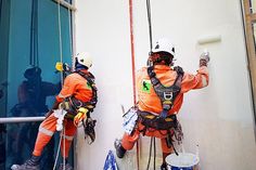 two men in orange safety gear climbing up the side of a building with ropes attached to them