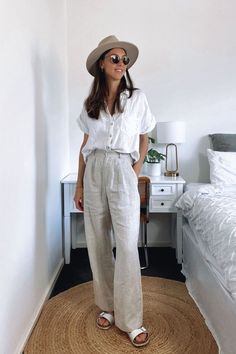 A girl wearing a neutral outfit featuring wide leg beige trousers, white birkinstock sandals, and a short sleeve linen shirt. She is also wearing a hat and sunglasses. Wide Leg Trousers Outfit Work, Summer Trousers Outfits, Trousers Outfit Work, Trousers Outfit Summer, Leg Trousers Outfit, Linen Trousers Outfit, Linen Pants Outfit Summer