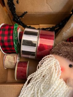 an assortment of christmas ribbons and decorations in a cardboard box with a doll next to them