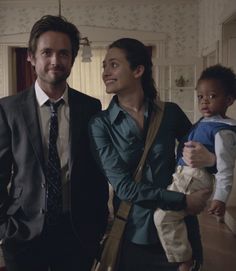 a man and woman standing next to each other in a living room with a baby on his lap