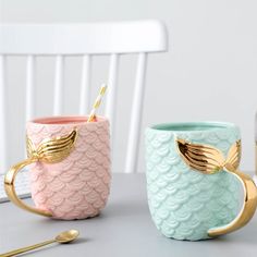 two mermaid mugs sitting on top of a table next to a fork and spoon