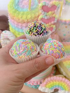 a hand holding three colorful cupcakes with sprinkles on them in front of a rainbow cake