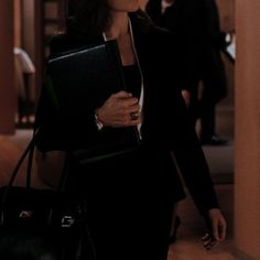 a woman in a suit carrying a black purse and looking at the camera while walking down a hallway