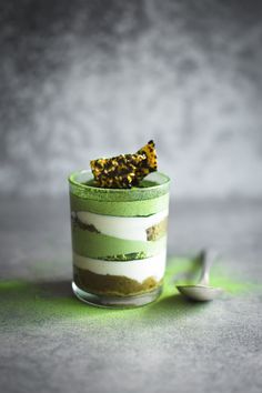 a green and white dessert in a small glass with a spoon next to it on a gray surface