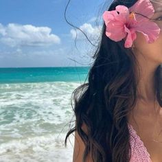 a woman standing on top of a beach next to the ocean with a pink flower in her hair