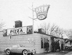 an old black and white photo of people standing in front of a pizza shop with a car parked outside