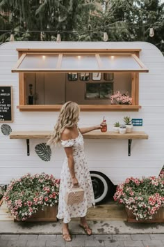 a woman standing in front of a food truck with flowers on the ground and plants behind her