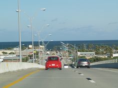 a red car is driving down the road by the ocean and street lamps are in front of it