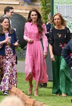 two women in pink dresses are walking through the grass with other people behind them and one woman is wearing green pants