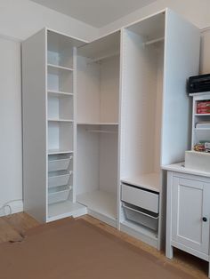 an empty white closet with shelves and drawers on the wall, next to a bed