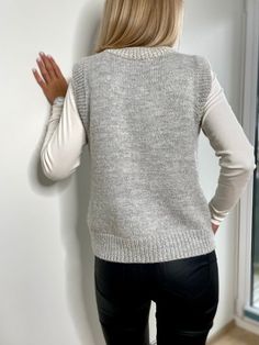 a woman standing against a wall with her back to the camera, wearing a gray sweater and black pants