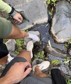 two people standing next to each other in front of some rocks and water with plants growing out of them