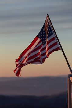 the american flag is waving in the wind on top of a building with mountains in the background