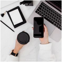 a person holding an iphone next to a laptop and other items on a white table