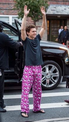 a man in purple pants standing next to a black car with his arms up and hands raised