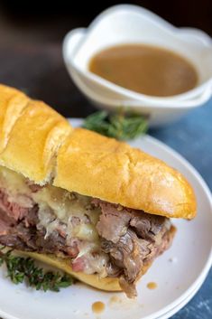 a roast beef sandwich on a white plate next to a cup of gravy