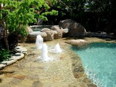 an outdoor pool with water jets and rocks in the middle, surrounded by greenery