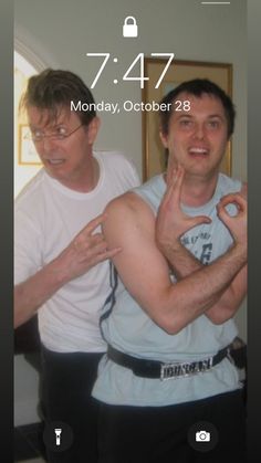 two men standing next to each other in front of a clock on a cell phone