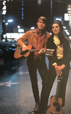 a man and woman standing next to each other holding guitars in the street at night