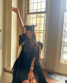 a woman in a graduation cap and gown is sitting on a window sill with her hand up