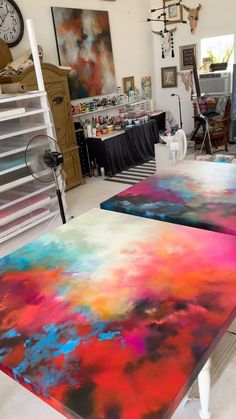 two tables painted with different colors in a room filled with art supplies and paintings on the walls